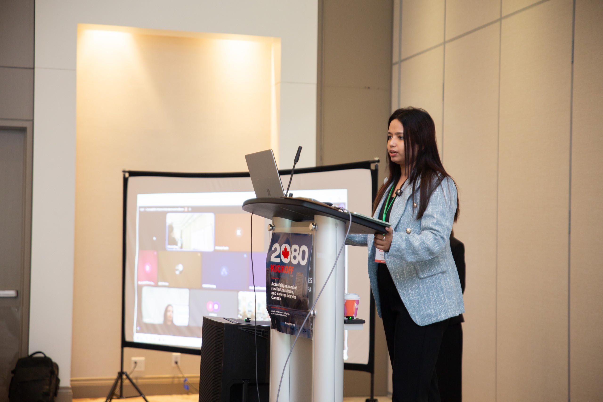 Sunita Kumari speaking at the podium in a light blue blazer, with remote participants on the screen behind her.
